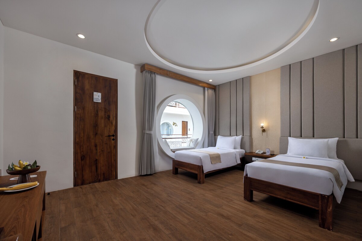 A hotel room with two beds, a wooden floor, a round mirror, a window with curtains, and a small table with a bowl of fruit.