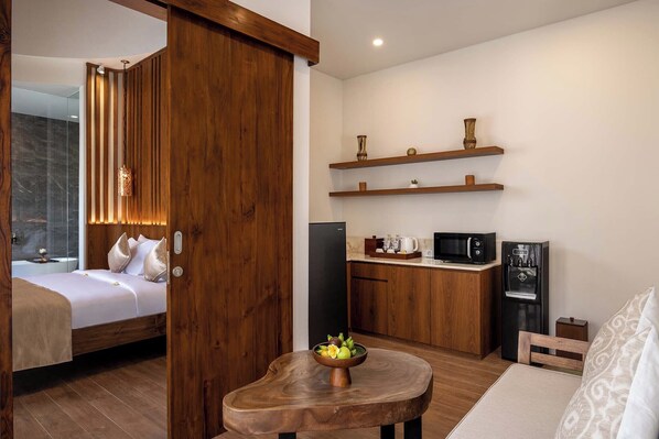 A compact hotel room with a wooden bed, a small kitchenette, and a dining area with a wooden table and bowl of fruit. - The Prime Canggu Bali (Canggu)
