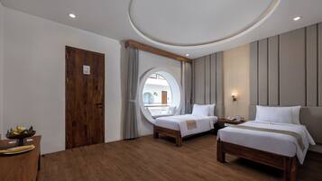 A hotel room with two beds, a wooden floor, a round mirror, a window with curtains, and a small table with a bowl of fruit.