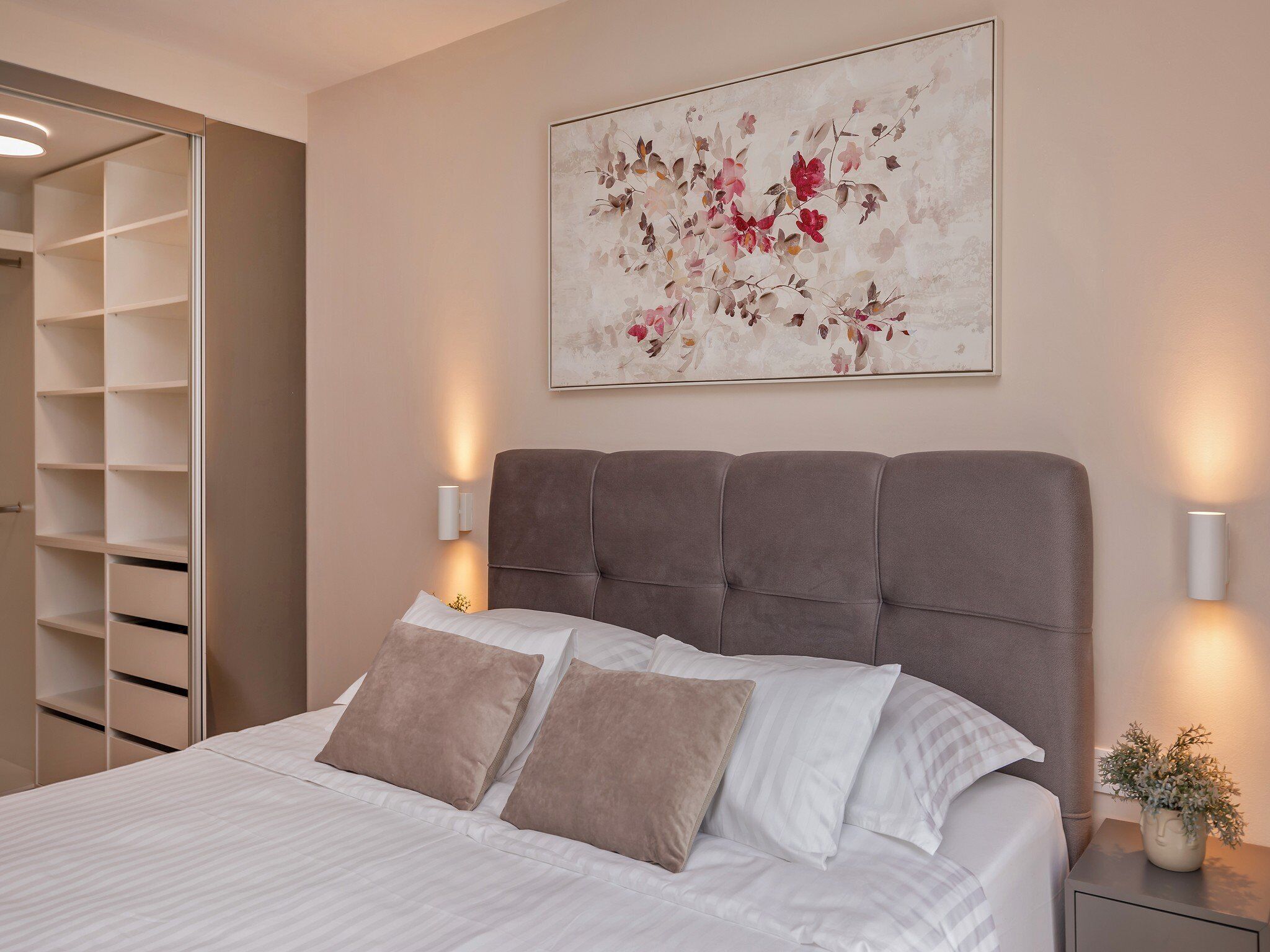 A bedroom with a bed, a nightstand, a wall-mounted light, a painting, and a closet.