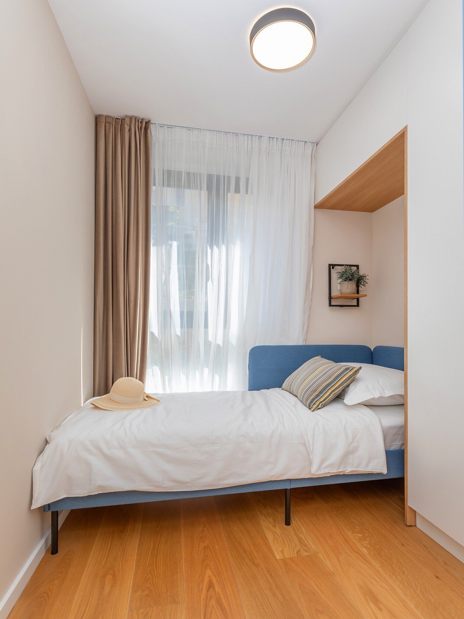 A small, neatly arranged bedroom with a bed, a window with sheer curtains, a small shelf with a plant, and a ceiling light.