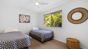 A bedroom with two beds, a ceiling fan, a window with a view of palm trees, a wall-mounted artwork, and a decorative circular wall piece.