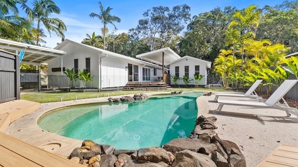 A pool area with a clear blue pool, surrounded by rocks and wooden decking. There are lounge chairs and a covered patio area with a view of the surrounding trees and buildings.