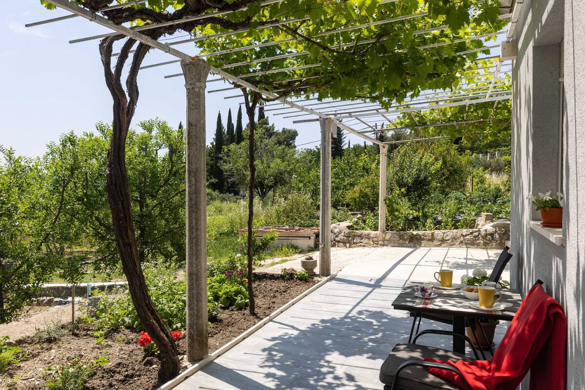 Un porche cubierto con una mesa y sillas, rodeado de un jardín con diversas plantas y una estructura de pérgola.