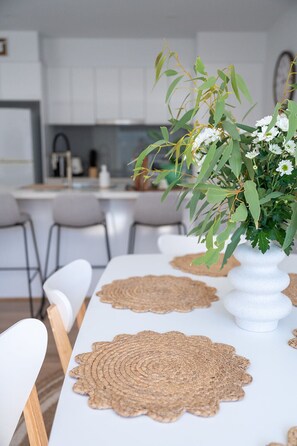 Mesa de jantar com jogos americanos trançados, um vaso branco e folhagem verde com flores brancas.