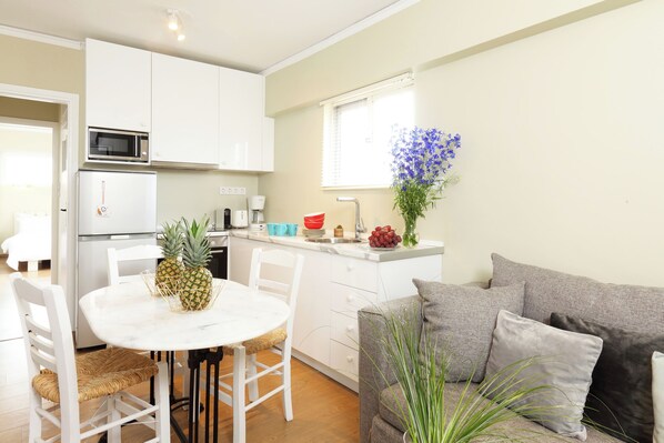 A compact kitchen with white cabinets, a microwave, and a refrigerator. There's a dining table with chairs and a small island with a sink. A grey sofa and a potted plant are in the living area. - Live in Athens, Our first love #01 (Athens)