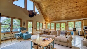A spacious living room with a high ceiling, large windows, a sofa, a coffee table, and a telescope. - Camp Muchemore! Hot Tub, Waterfront! (Hancock)