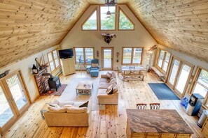 A spacious loft with a high ceiling, wooden floors, a large sofa, a dining table, and a fireplace. - Camp Muchemore! Hot Tub, Waterfront! (Hancock)