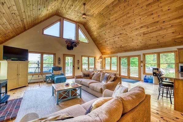 A spacious loft-style room with a high ceiling, large windows, a television, a sofa, a dining table, and a kitchen area. - Camp Muchemore! Hot Tub, Waterfront! (Hancock)