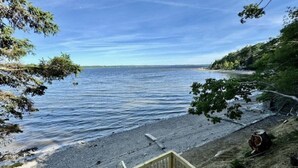 Ett trädäck med utsikt över ett lugnt vattenområde, en sandstrand och ett skogsområde.