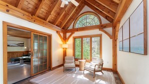 A spacious room with wooden beams, a large window, a fireplace, and a view of the outdoors. - New! Acadia Nature Escape w/Trails + Water Access (Sullivan)