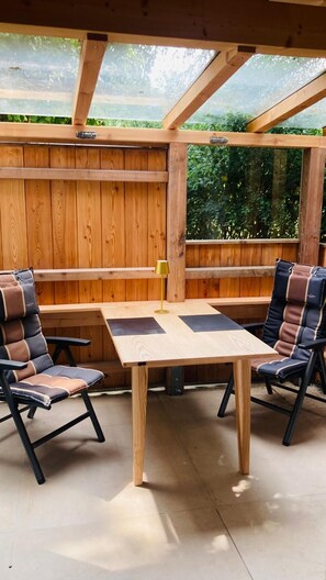Een houten terras buiten, met een tafel en stoelen, omgeven door houten wanden en een glazen dak.