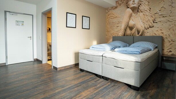 A bedroom with a bed, a wardrobe, a door, and a wall with a textured mural. - Apartment with shower (Garbsen)