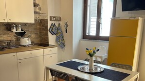 Uma cozinha compacta com armários brancos, piso ladrilhado, mesa de jantar com cadeiras e uma janela com persianas.