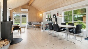 Un interno moderno con soffitto in legno, un caminetto, un'area pranzo con sedie, un divano e una grande finestra con vista sul verde.
