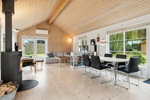 Et moderne interiør med træloft, pejs, spiseplads med stole, sofa og stort vindue med udsigt til grøn natur.