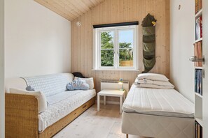 Et værelse med en fletsofa, et lille natbord med en lampe, en enkelt seng, et vindue med udsigt til grønne omgivelser og en bogreol.