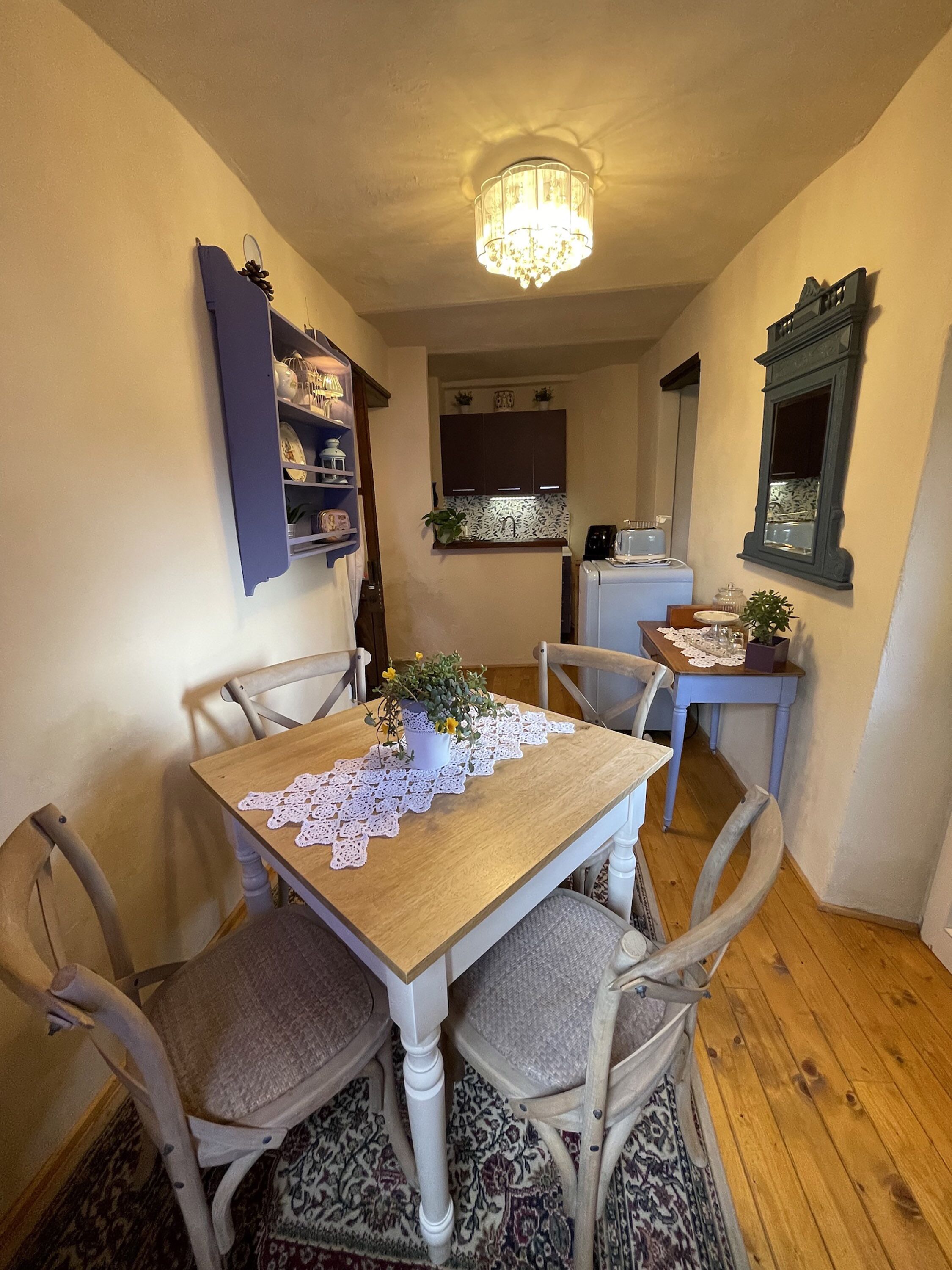 A dining area with a wooden table, four chairs, a chandelier, and a kitchen in the background.