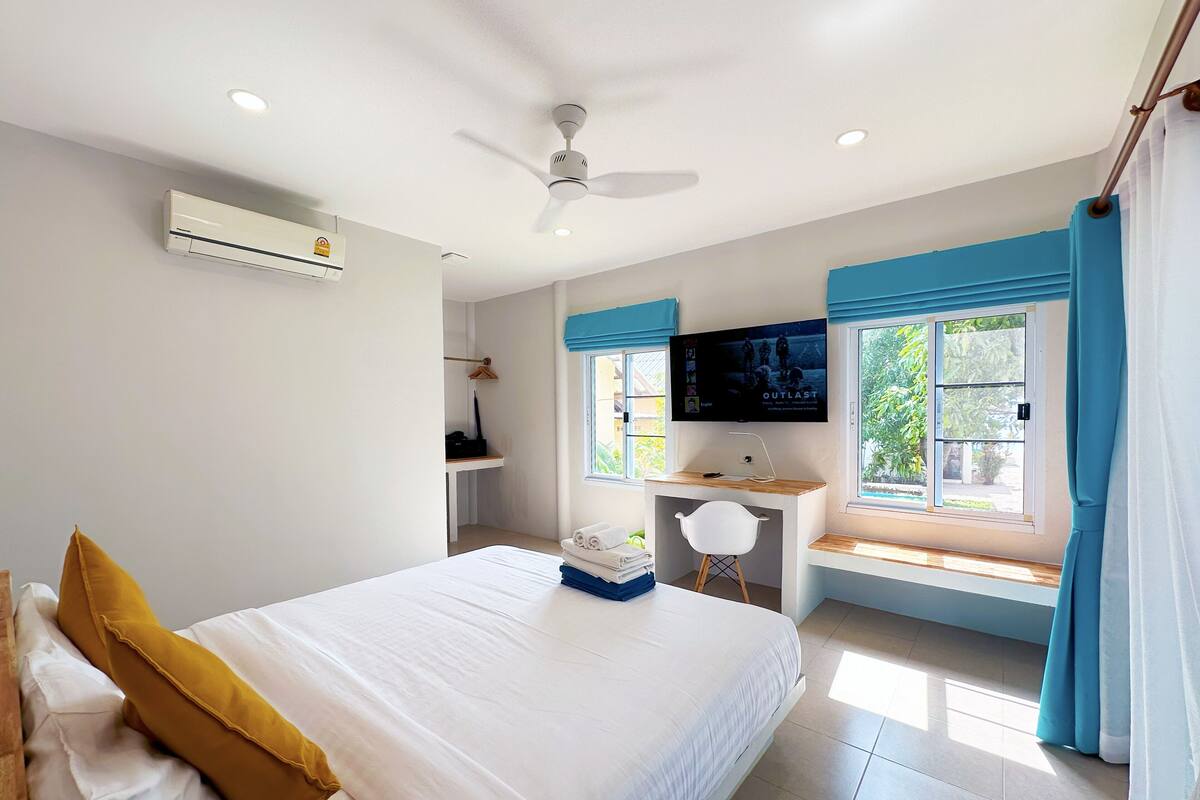 A bedroom with a bed, a desk, a chair, a TV, and a window with curtains.