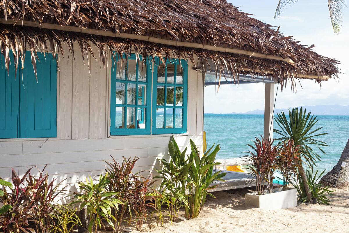 A beach house with a thatched roof, turquoise shutters, and a view of the ocean.
