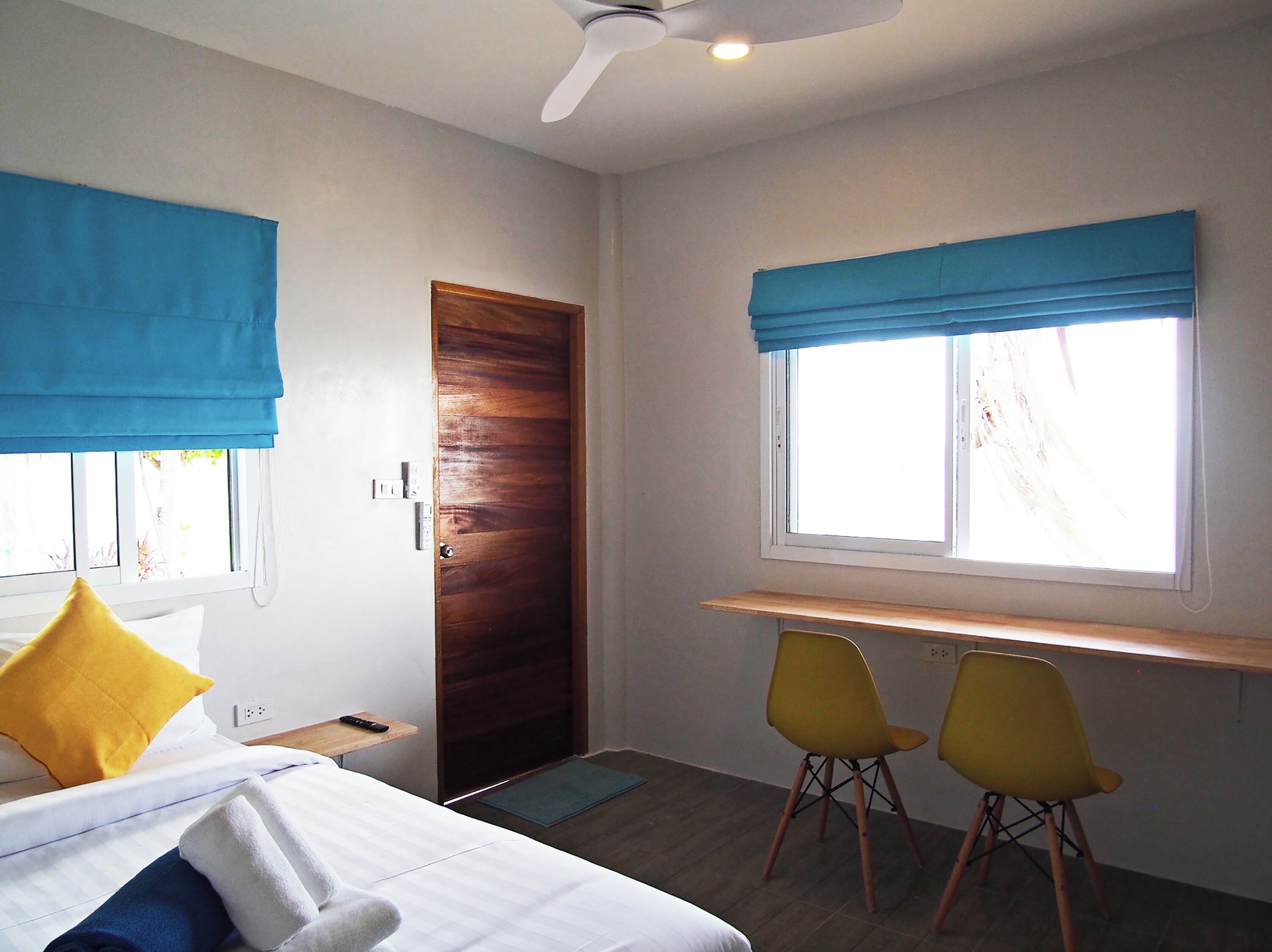 A bedroom with a bed, a desk, two chairs, a window with blue curtains, and a door.