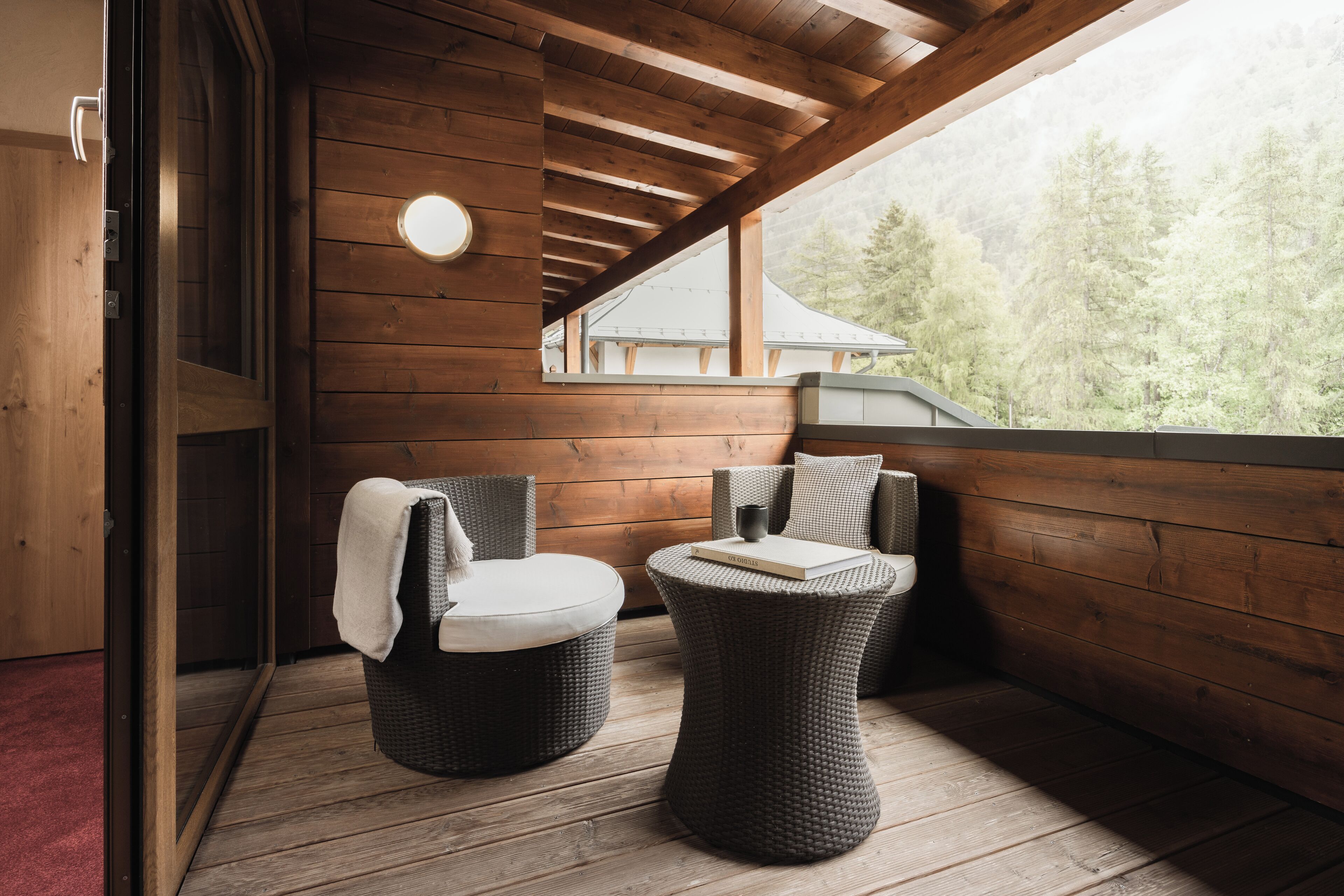 Un balcón de madera con dos sillones de mimbre, una mesa redonda y vistas a una casa y árboles.