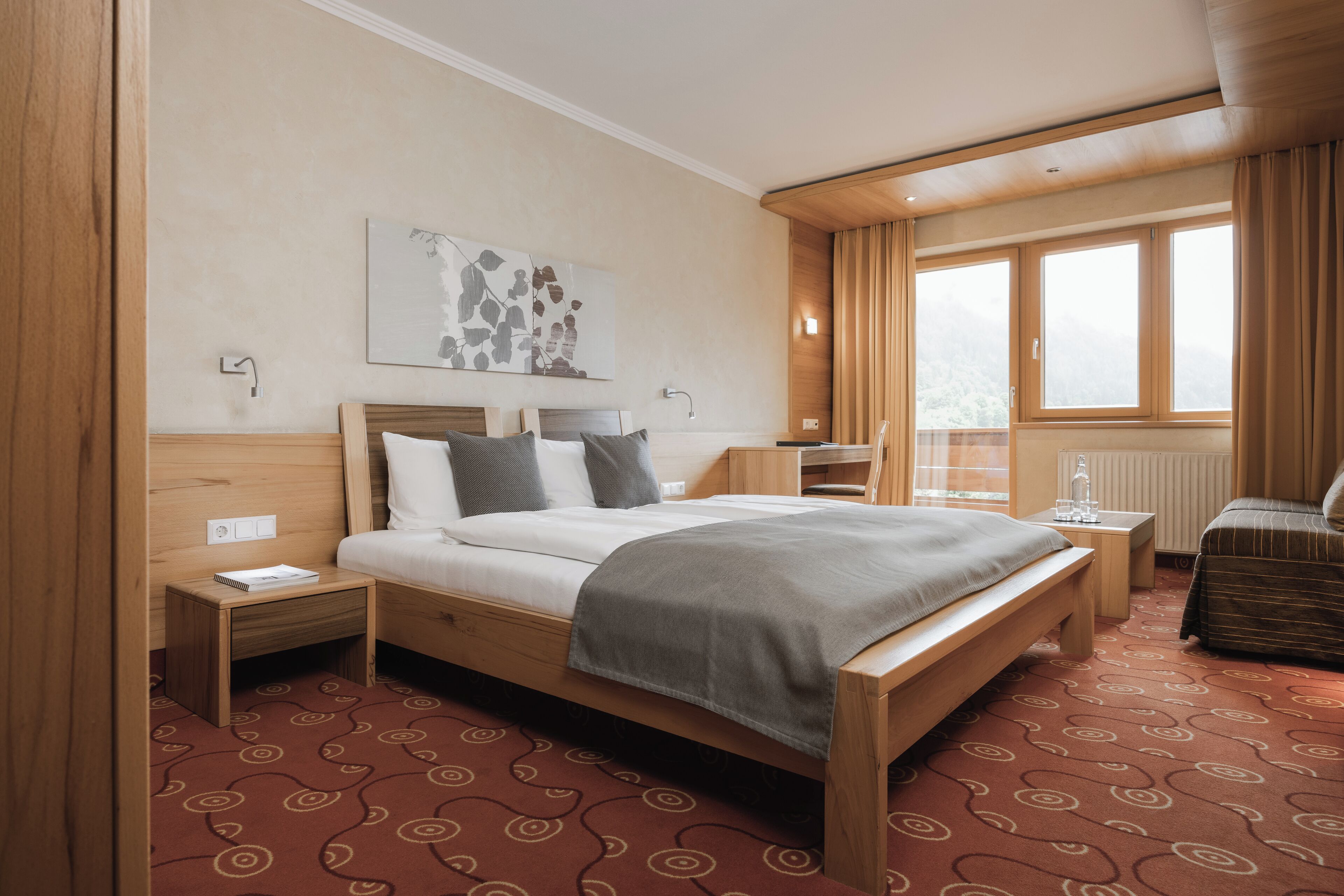 A hotel room with a bed, bedside tables, a nightstand, a chair, a window with curtains, and a wall-mounted artwork.