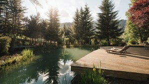 A wooden deck with two lounge chairs overlooking a calm pond, surrounded by dense trees and a mountain backdrop. - miaflor Familien- & Aktivresort (Ried im Oberinntal)