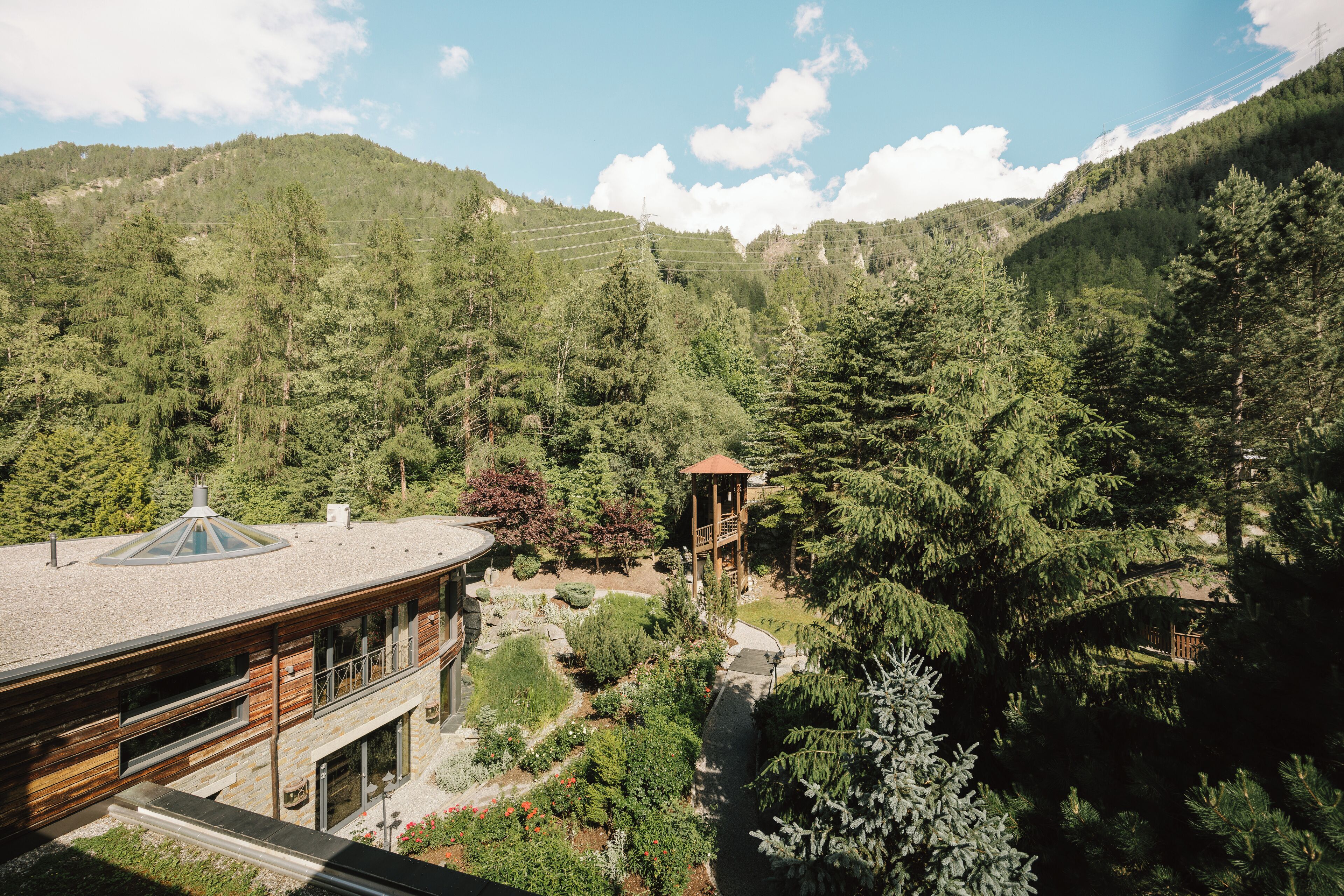 A modern building with a glass conservatory, surrounded by a lush garden and forested hills.