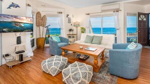 A living room with a wooden coffee table, two blue armchairs, a white sofa, and a view of the sea through large windows.