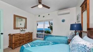 A room with two beds, a ceiling fan, a window with a view of the ocean, a wooden dresser, and a mirror.