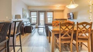 Un salon moderne avec un canapé, une table à manger et des chaises. Un téléviseur est fixé au mur et il y a une fenêtre avec des stores.