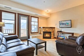 A living room with leather sofas, a fireplace, and a television displaying 'Stay In' on the wall. - Mtn Views + Bunk Room / Pool, Hot Tub, Sleeps 6 (Fernie)