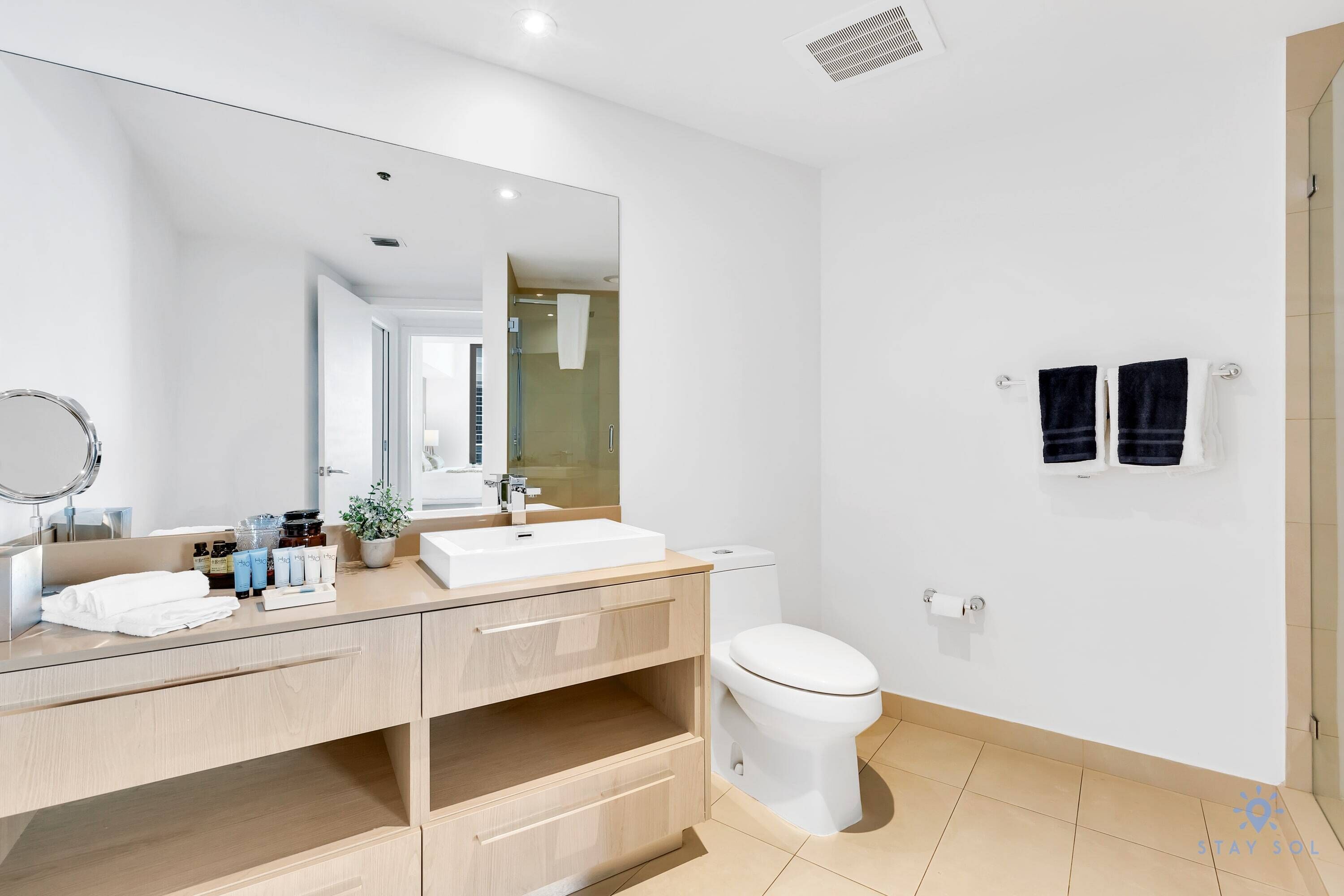 A modern bathroom with a large mirror, white sink, and toilet. There are two towels hanging on hooks and a potted plant on the vanity.