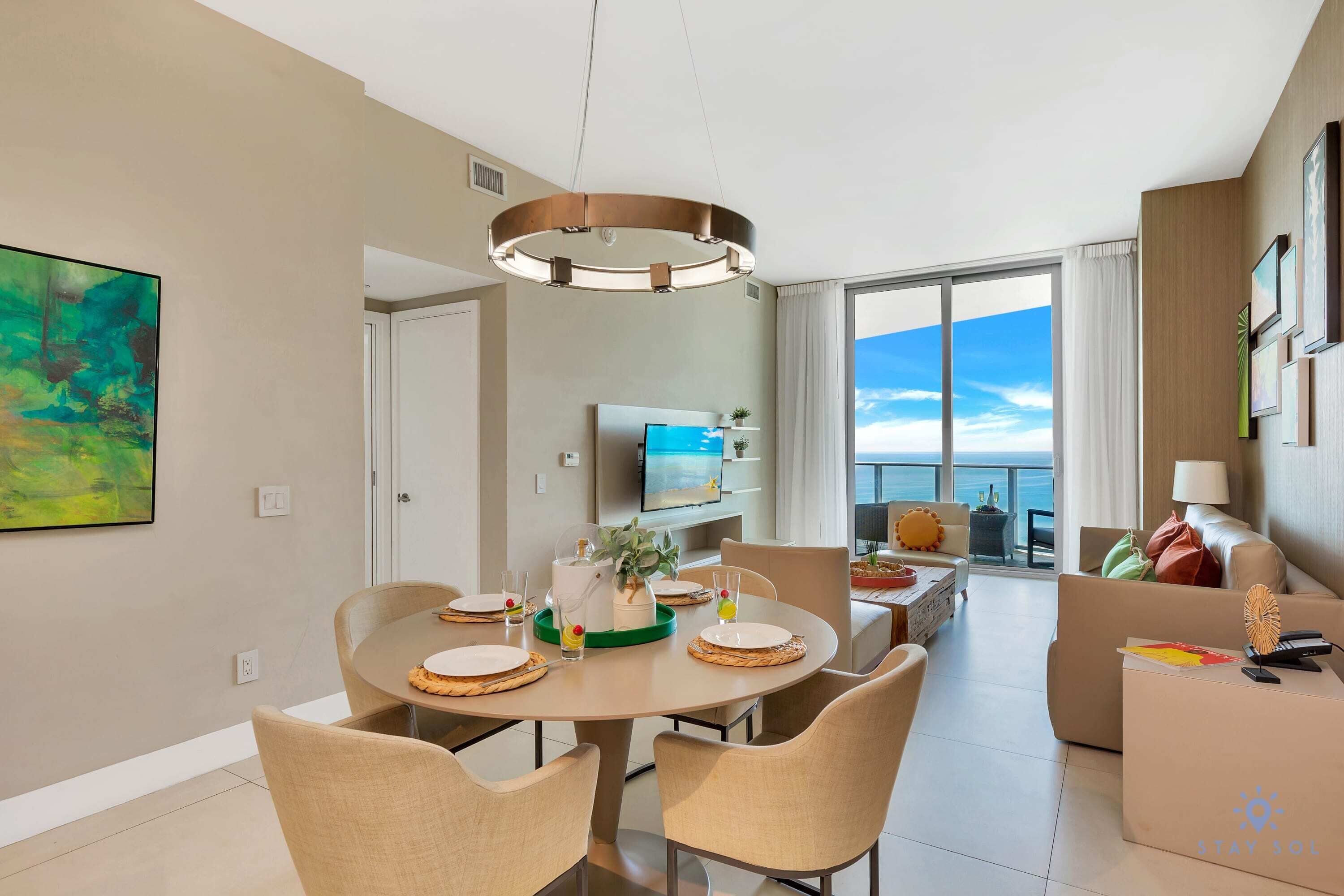 A modern hotel room with a dining area, a living area with a view of the sea, and a balcony with a large window.