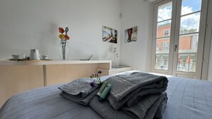 Uma cama bem arrumada com um cobertor cinza, uma mesa de cabeceira com um vaso de flores e uma janela com vista para edifícios.
