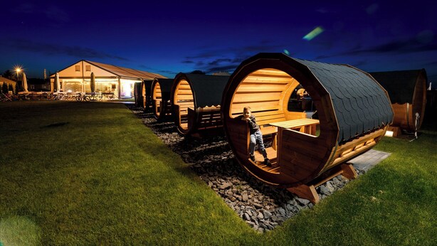 Une rangée de structures en bois en forme de tonneau, dont l’une est occupée par une personne, et qui sont reliées à un bâtiment dont l’entrée est illuminée.
