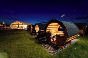 A row of barrel-shaped wooden structures with a person inside one, connected to a building with a lit entrance. - Fantazja Resort Zator (Zator)
