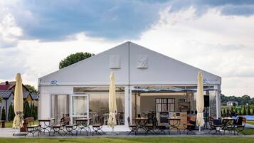 A white pavilion with a flat roof, large glass windows, and a covered outdoor seating area with tables and chairs.