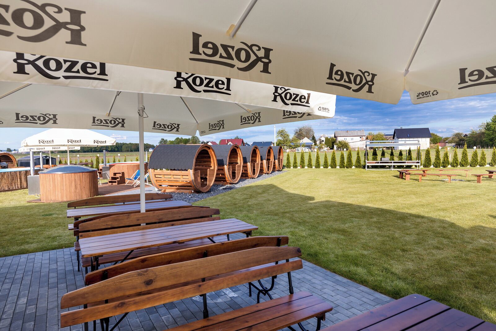 Un área de estar al aire libre con bancos y mesas de madera bajo grandes sombrillas blancas con el logo de 'Kozel'.