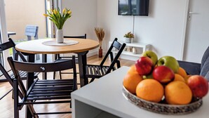 Un coin repas avec une table ronde en bois, des chaises noires, un vase avec des fleurs jaunes, un saladier rempli de pommes et d’oranges, un téléviseur à écran plat fixé au mur et une fenêtre donnant sur l’extérieur.