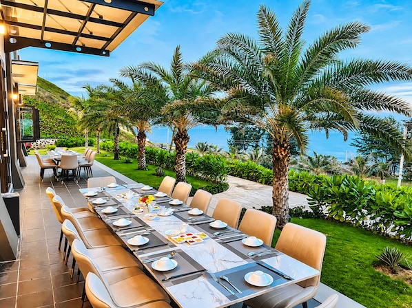 An outdoor dining area with a long table set for a meal, surrounded by palm trees and a view of the ocean.
