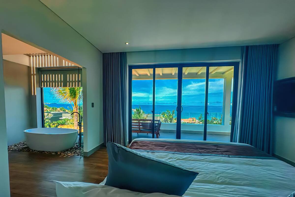 A modern bedroom with a large bed, a bathtub, and a balcony with a view of the ocean.