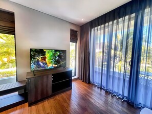A modern living room with a flat-screen TV on a wooden stand, large windows with sheer curtains, and a wooden floor. - Du Parc Resort Mũi Né (Phan Thiet)