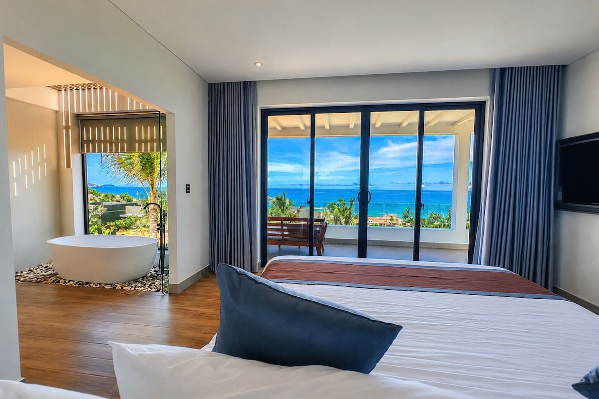 A modern bedroom with a large bed, a bathtub, and a balcony with a view of the ocean.