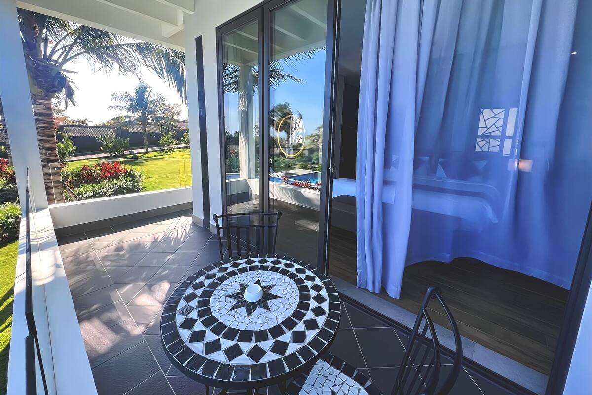 A modern outdoor patio with a mosaic table, chairs, and a view of a pool and garden.