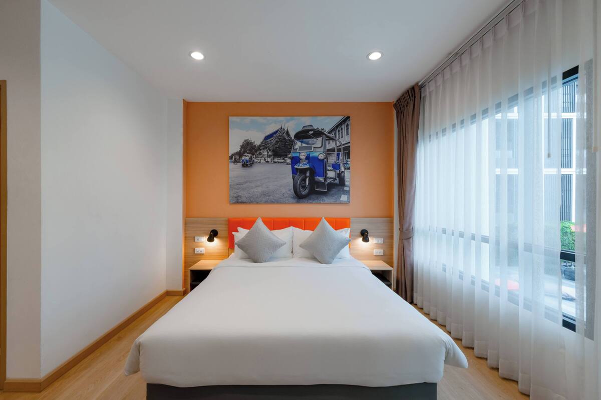Une chambre d’hôtel avec un grand lit, deux tables de chevet avec des lampes, un tableau au mur et une fenêtre avec des rideaux légers.