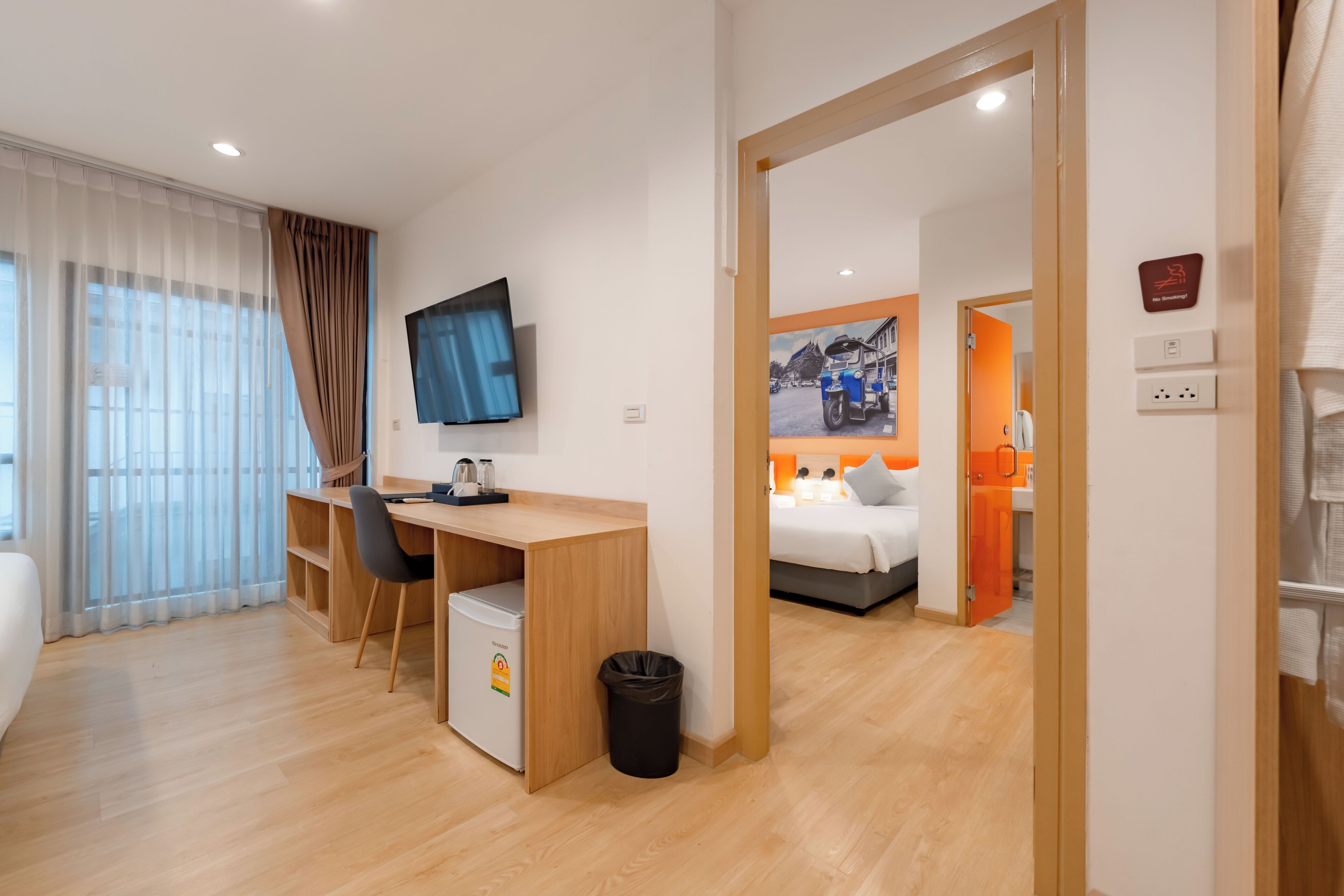 A modern hotel room with a wooden desk, a small refrigerator, a TV, and a bed with an orange accent wall.