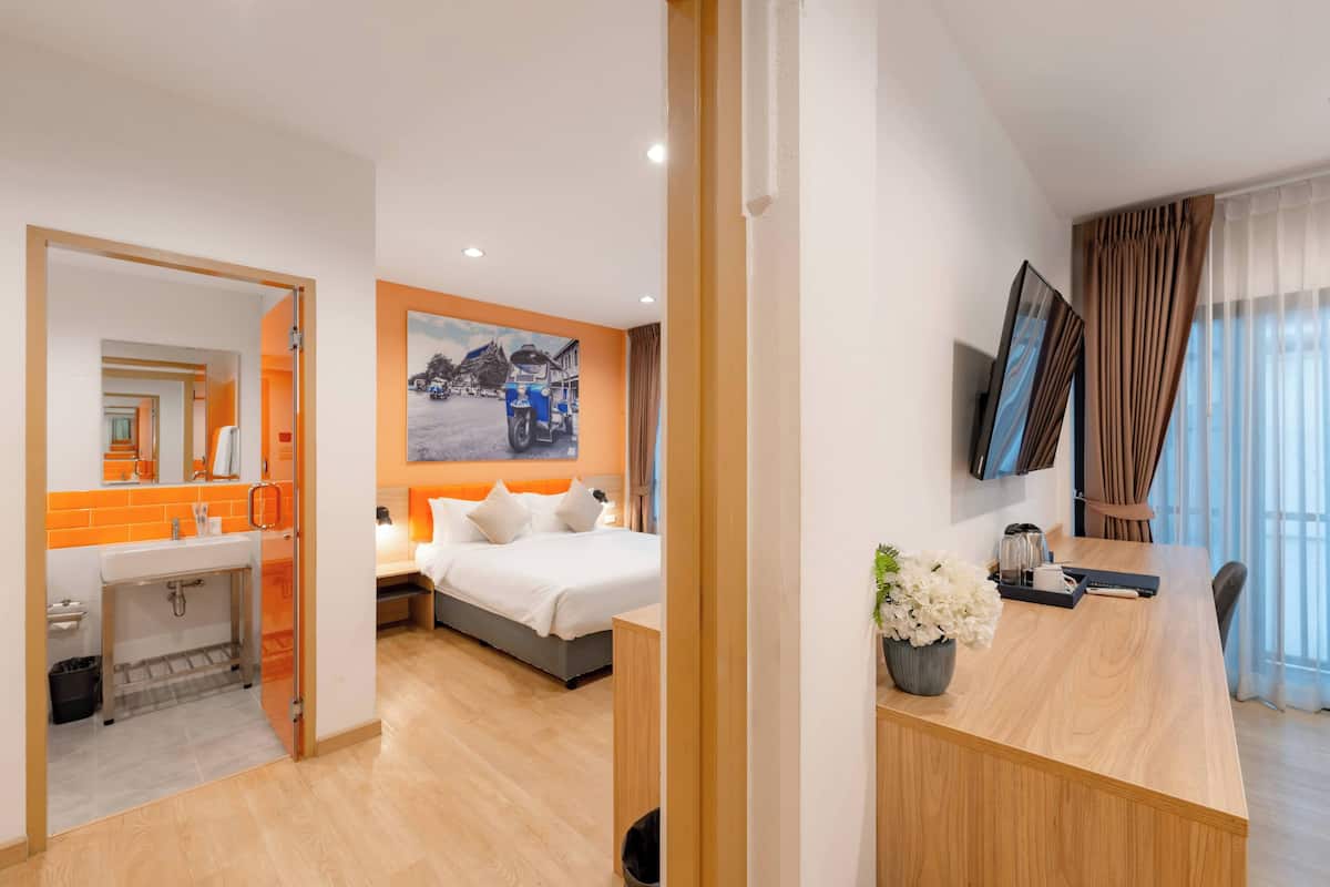 Une chambre d’hôtel avec un lit, un bureau agrémenté d’une plante, une télévision et une salle de bain dont un mur est peint dans une teinte orange.