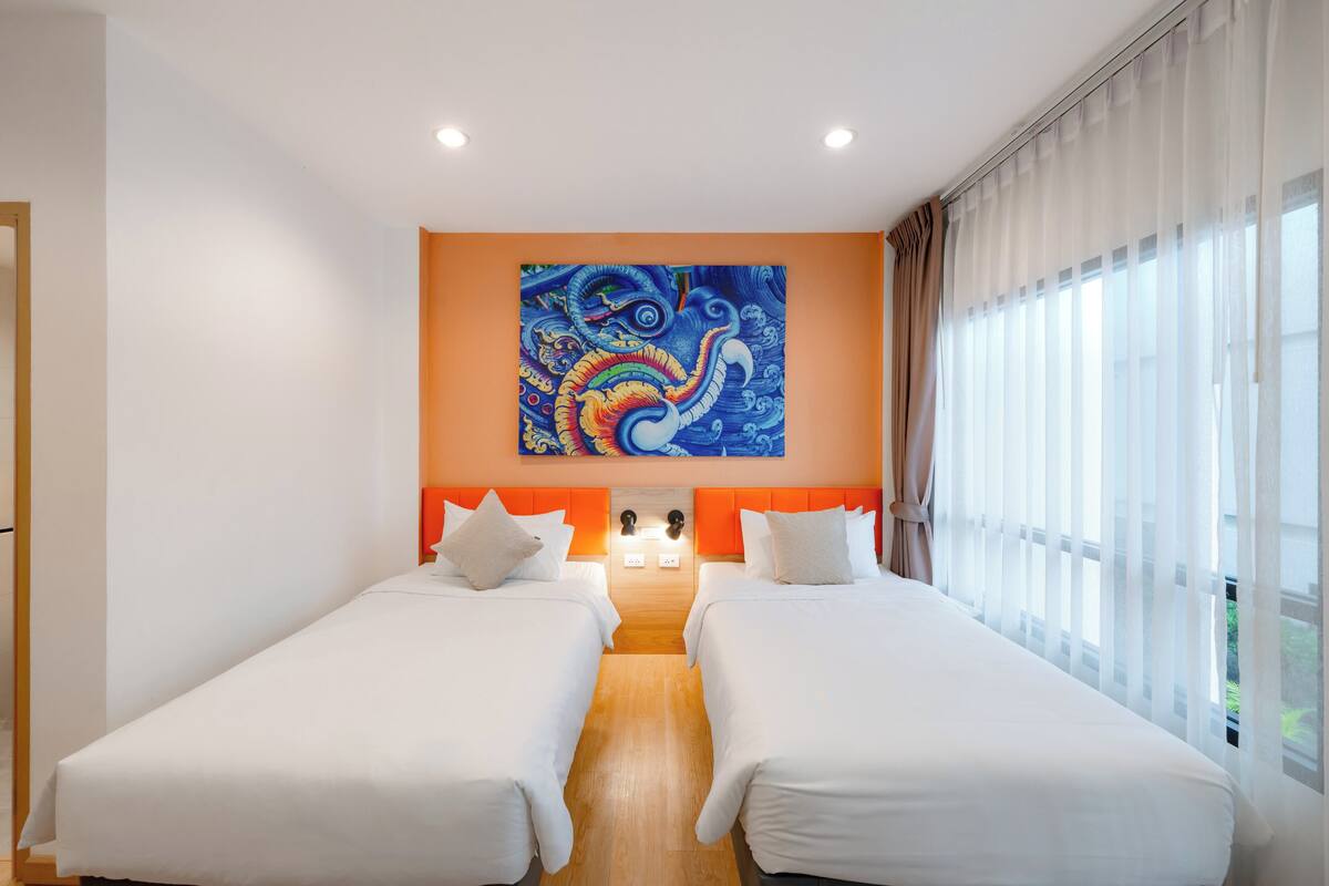 Une chambre d’hôtel avec deux lits, une grande fenêtre ornée de rideaux légers et une peinture abstraite aux couleurs vives accrochée au mur.